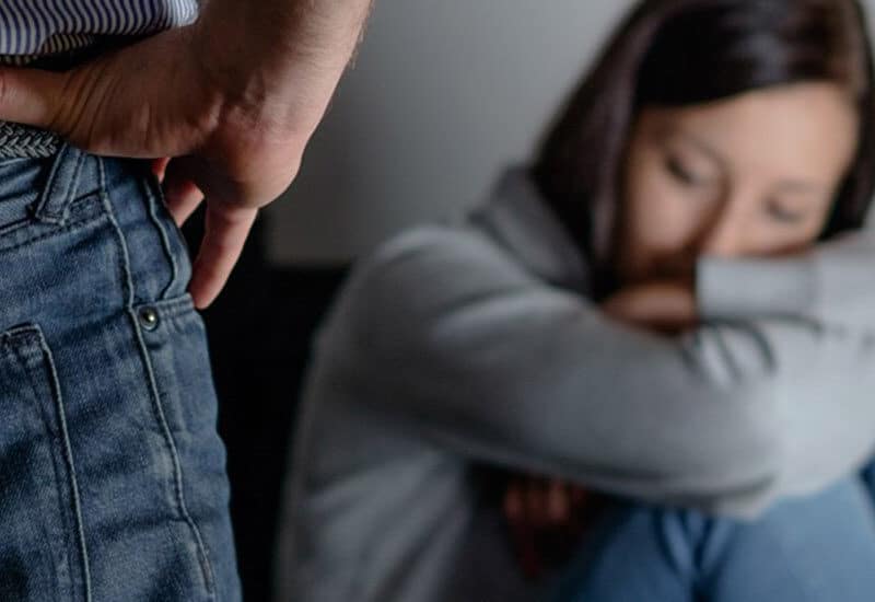 cropped image of man with hands on hip standing in front of woman sitting on floor curled up with head down due to domestic violence